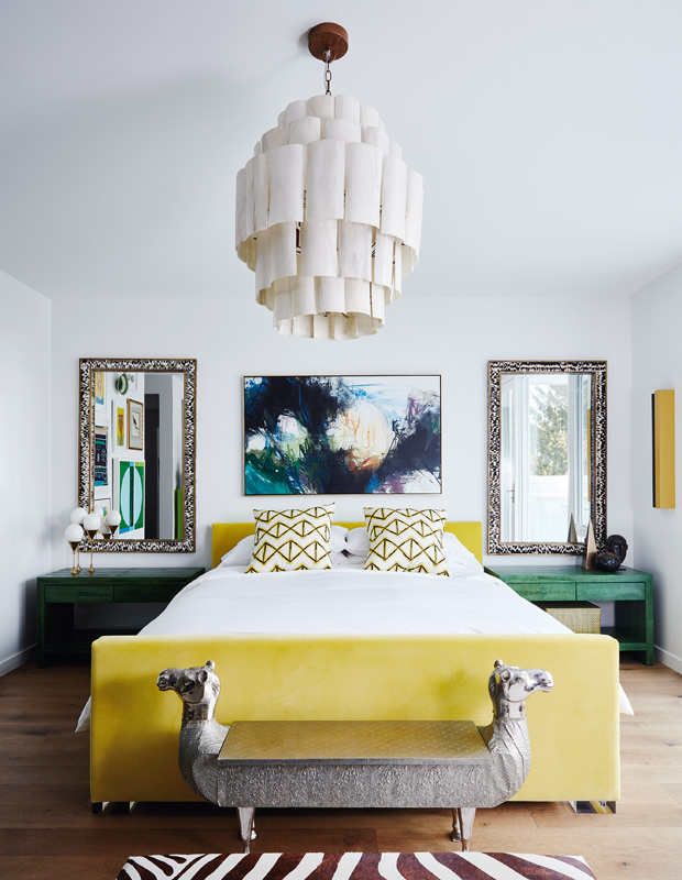 A stylish maximalist bedroom done in white, with a yellow upholstered bed, green nightstands, mirrors, a statement artwork and a catchy chandelier