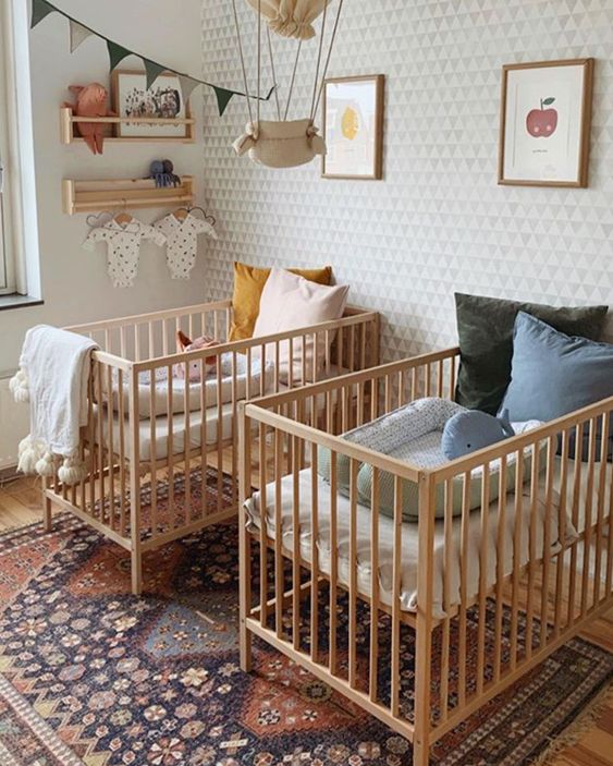 a stylish mid-century modern twin nursery with a wallpaper wall, stained cribs, a printed rug and pastel and printed bedding plus art