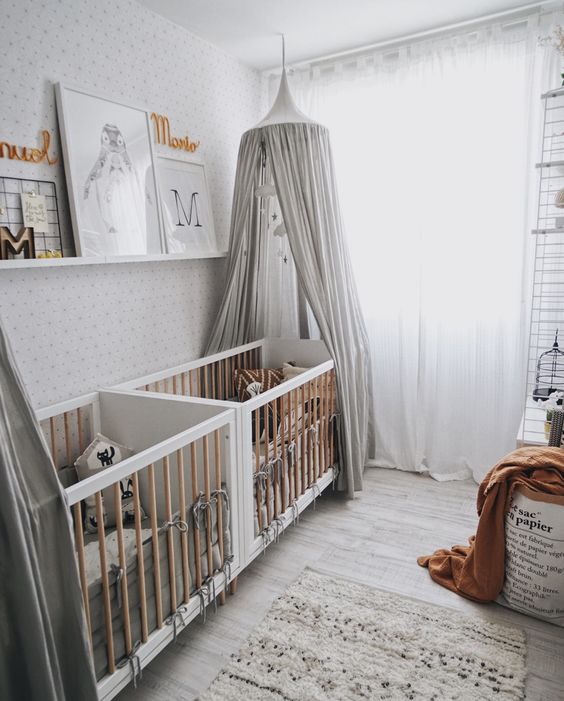 a stylish neutral nursery with white cribs, a rug, grey canopies, a ledge with artworks and lovely textiles