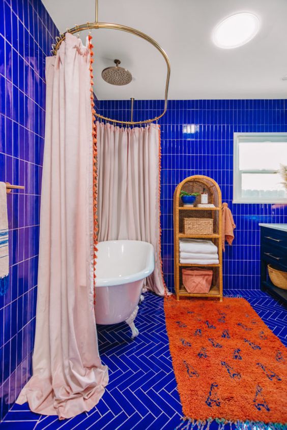 a super vibrant maximalist bathroom clad with electric blue tiles, a vintage tub with a large curtain, an orange rug, a rattan shelf and a black vanity