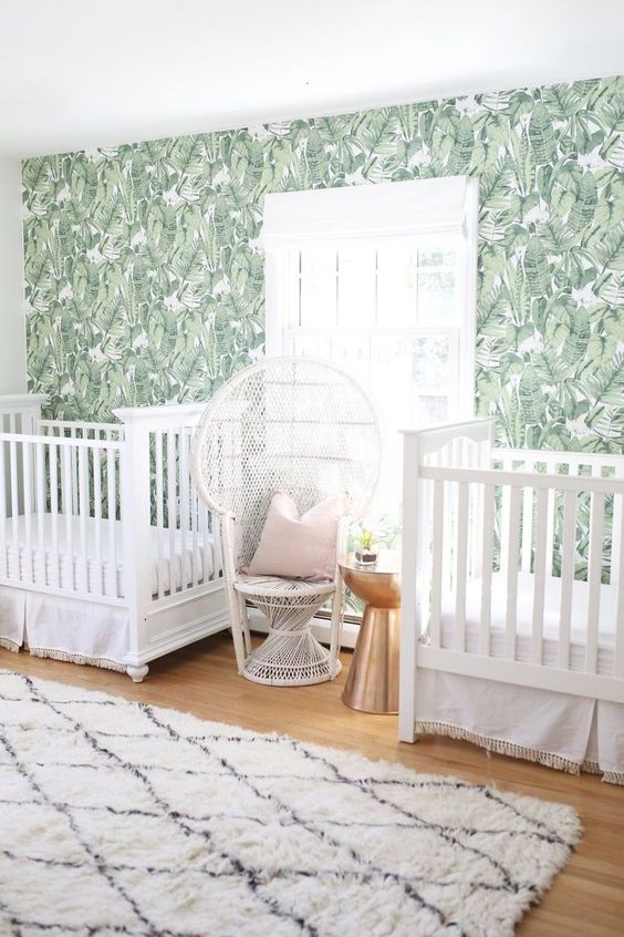 a tropical twin nursery with a banana leaf wall, white cribs and a peacock chair, a printed rug and a metallic side table