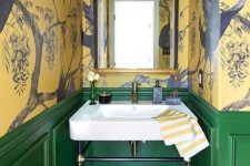 a vintage maximalist bathroom with yellow bird wallpaper walls, green paneling, a black hex tile floor, a mirror in a gilded frame and a lamp