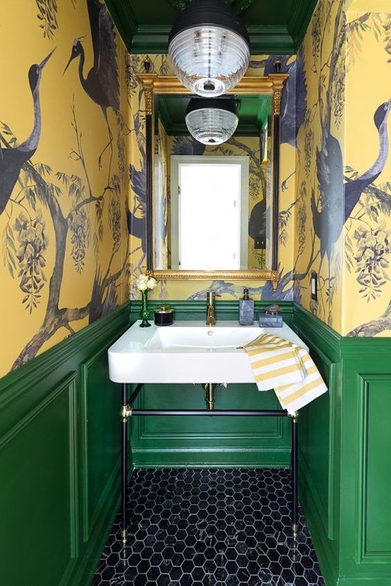 a vintage maximalist bathroom with yellow bird wallpaper walls, green paneling, a black hex tile floor, a mirror in a gilded frame and a lamp
