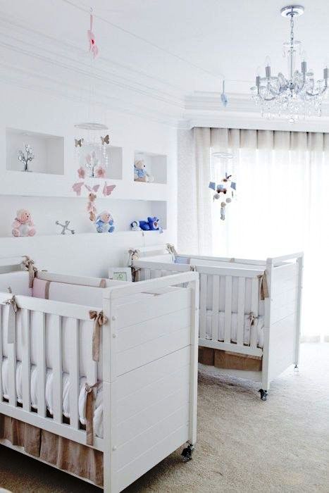 A white twin nursery with pretty cribs, built in shelves, a crystal chandelier and some pastel and bright toys is cool