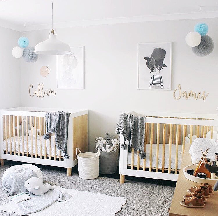 an airy contemporary nursery with two matching cribs, neutral textiles and layered rugs, lovely artworks and pendant pompoms