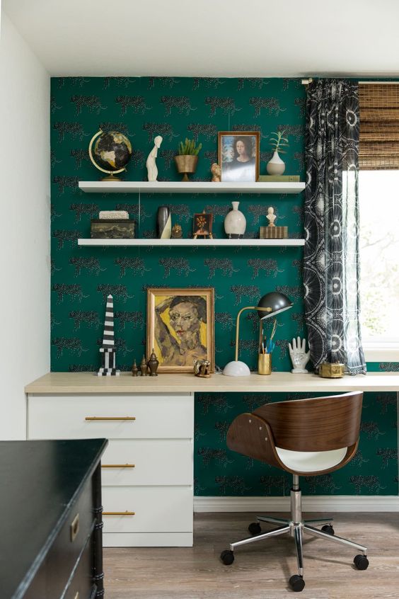 an elegant maximalist home office with a green wallpaper wall, a large desk, a comfy chair, a black dresser, lots of accessories and printed curtains