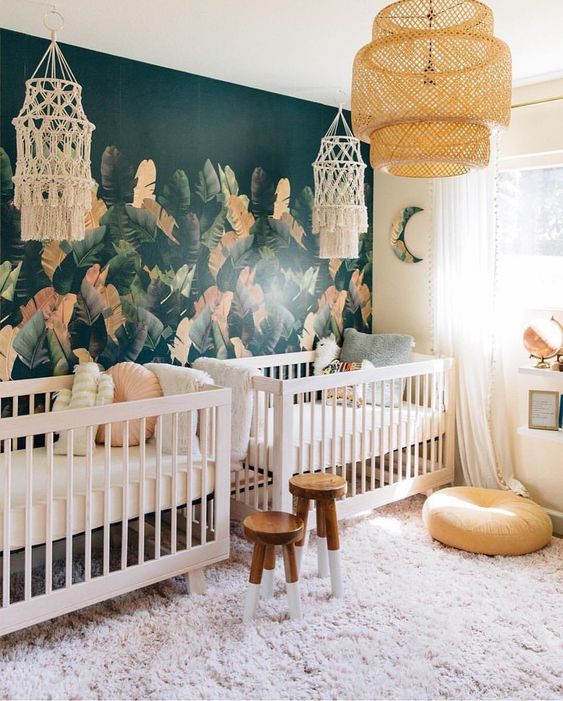 An eye catchy boho twin nursery with a tropical leaf accent wall, white cribs, wooden stools, macrame and a pendant lamp is wow