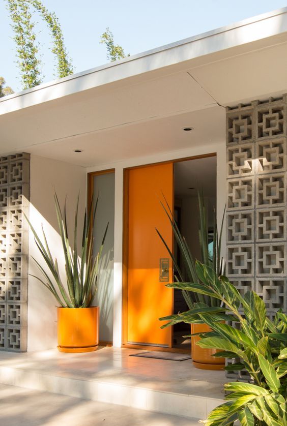 A very chic mid century modern front porch with a bold orange door and planters with oversized plants plus screens to protect from the sun