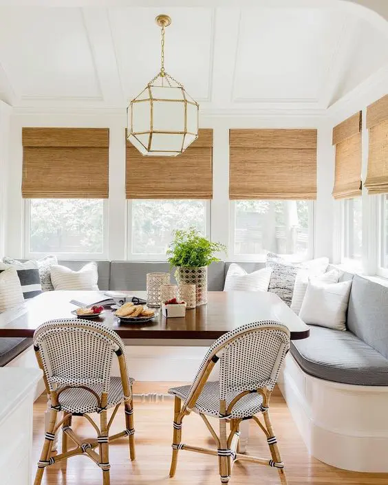 a chic and lovely dining space with corner windows, a large upholstered banquette seating, a dining table and rattan chairs