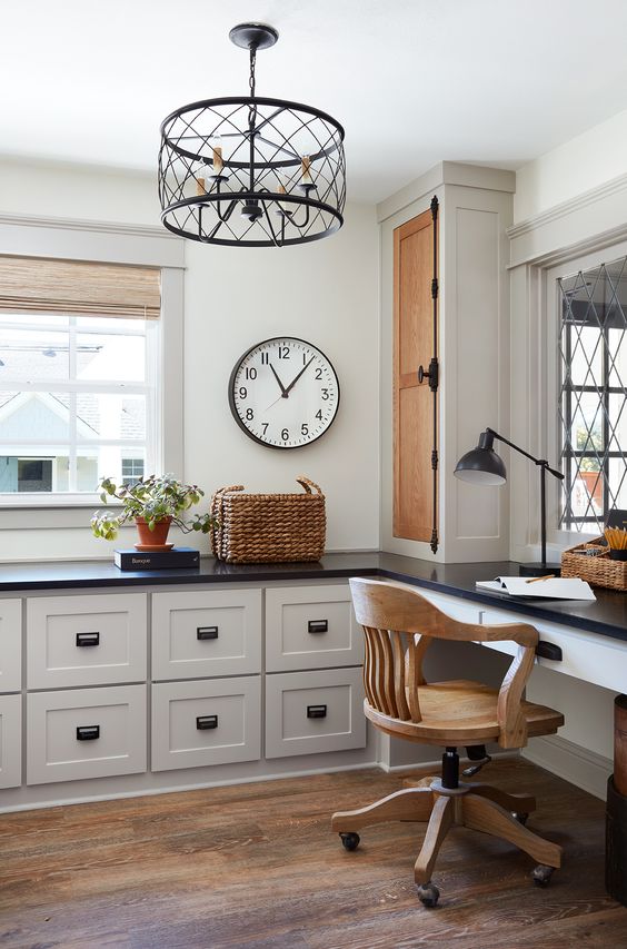 a stylish farmhouse home office done with pretty kitchen cabintery and a built-in desk, a wooden chair, a metal chandelier and a basket
