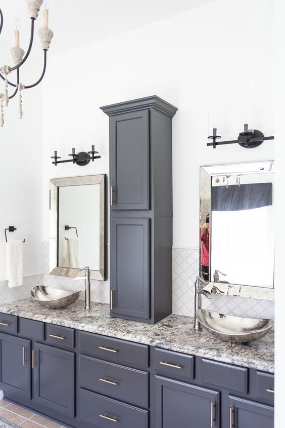 a midnight blue bathroom done with various kitchen cabinets and a grey stone countertop, hammered bowl sinks and a vintage chandelier