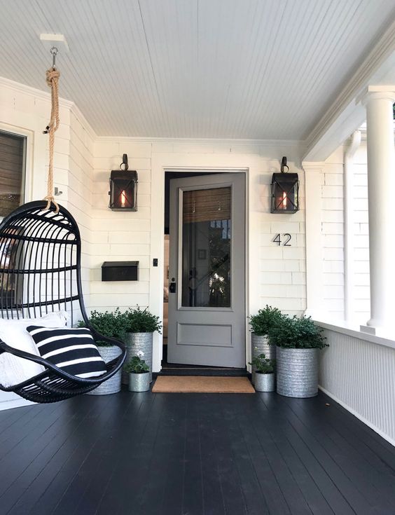 A modern porch with a black planked floor, a black pendant egg shaped chair, lots of greenery in tin cans and lanterns on the wall