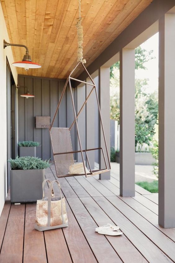 a modern porch with pillars, a planked wooden floor, a hanging metal chair, metal planters with greenery looks ultimate chic