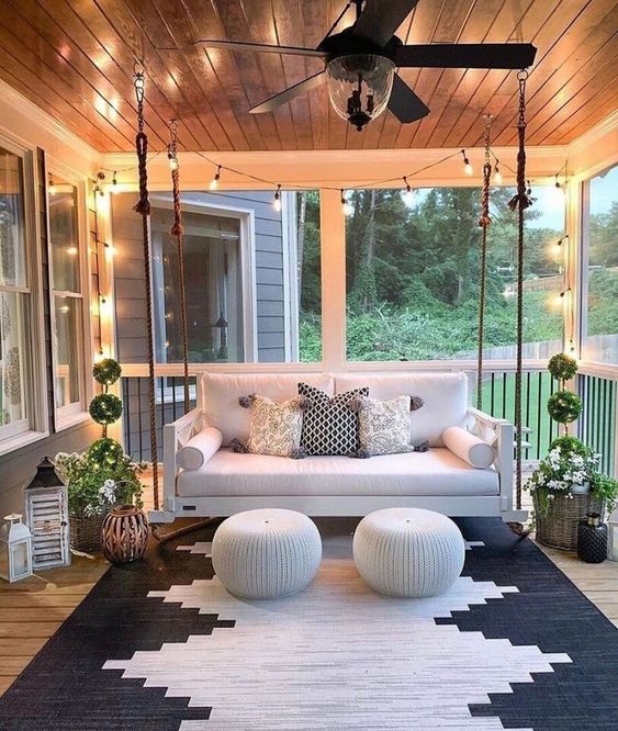 a welcoming modern porch with a suspended sofa, matching poufs, potted plants, candle lanterns and a bold geometric rug plus lights over the space