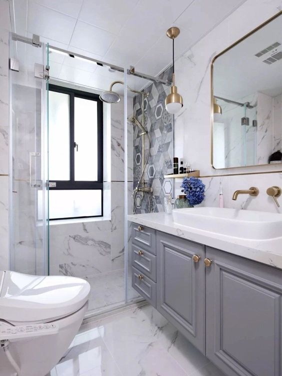 a beautiful fancy bathroom with white marble, a mosaic tile wall in the shower, a grey vanity, gold touches here and there