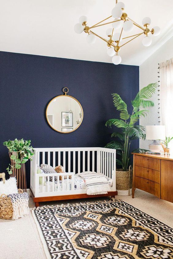 a beautiful modern nursery with a navy accent wall, chic mid-century modern furniture, a printed rug, a basket for storage, potted plants and a chic chandelier