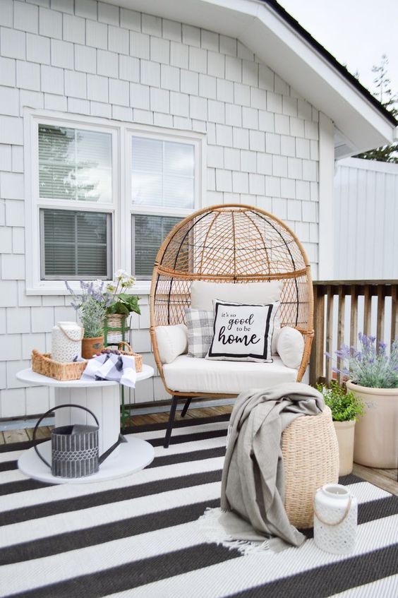 a boho farmhouse patio with a striped rug, a wicker egg-shaped chair, some baksets, potted blooms and a rustic side table with blooms
