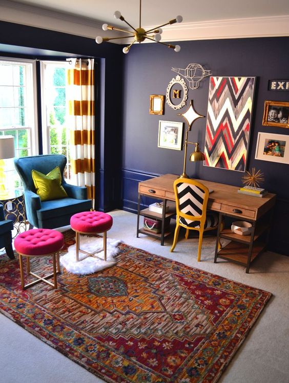 a bold home office with navy walls, a wooden desk, a blue chair and hot pink stools and a bold gallery wall