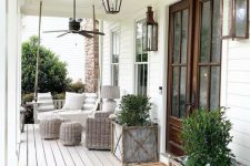 a charming farmhouse porch with a hanging bench with striped pillows, a wicker chair and poufs, plants in wooden boxes and rustic vintage lanterns