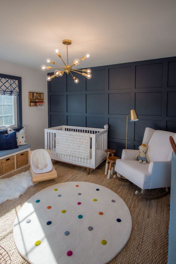 a cheerful nursery with a navy paneled wall, white furniture, a bench with storage, layered rugs and a cool modern chandelier