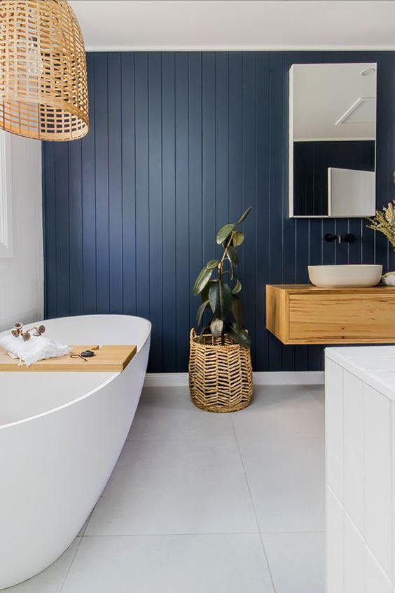 a chic coastal farmhouse bathroom with a navy planked wall, a large tub with a cuddy, a wooden pendant lamp and a floating vanity plus a cool mirror