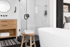 a chic fancy bathroom with a black hex tile floor, an oval tub, wooden furniture and a striped rug plus a glass enclosed shower