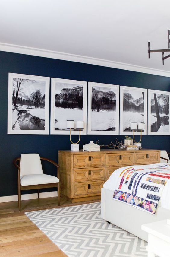 a chic farmhouse bedroom with a navy accent wall, a black and white gallery wall, a stained dresser with inlays, a comfy bed and pretty chairs