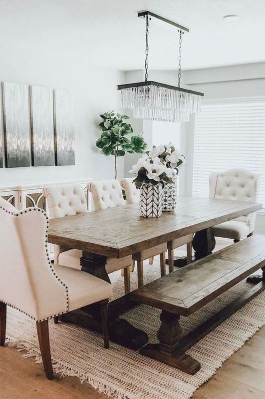 a chic modern country dining room with white walls, a vintage wooden table and a bench, tufted chairs and a lovely crystal chandelier