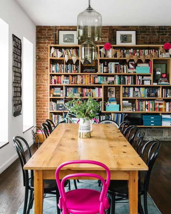 a cool industrial space with an extended brick wall, a wooden table, a bookshelf that takes a whole wlal and a hot pink chair for an accent