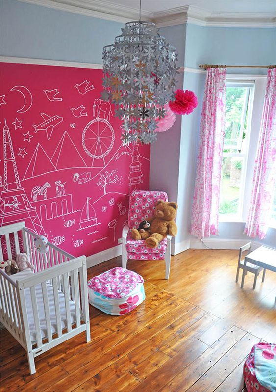a cool kid's room with a hot pink accent wall, a crib and a comfy chair with a pouf, a white dining space and a floral chandelier