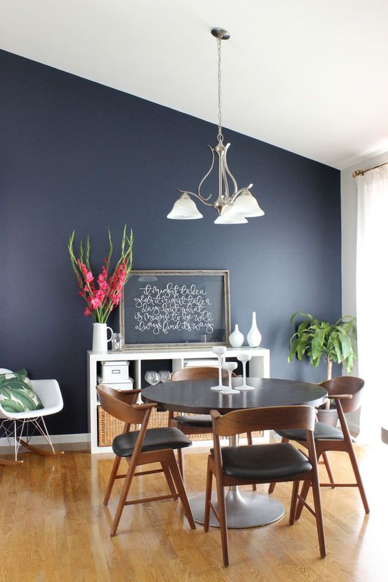 a cool modern dining room with a navy accent wall, a round table and black chairs, a storage unit, greenery and blooms