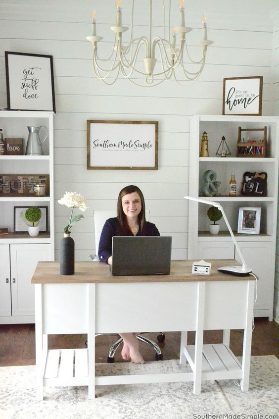 A cute white modern country home office with a planked wall, built in storage units, a large desk and a neutral chandelier