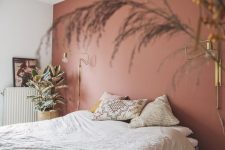 a dreamy and relaxed bedroom with a dusty pink accent wall, a bed and printed pillows, some potted plants and gilded sconces