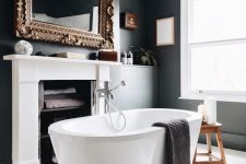 a fancy bathroom with black walls and a white planked floor, a fireplace used for storign towels, an oval tub and a mirror in a refined frame