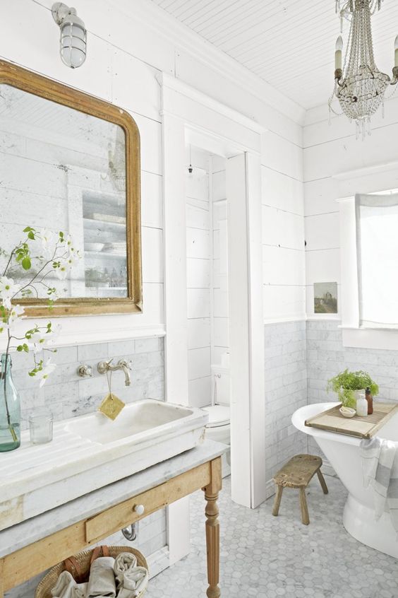 a fancy bathroom with grey marble tiles, planked walls, a vintage wooden vanity and a mirror in a wooden frame, a crystal chandelier and potted greenery
