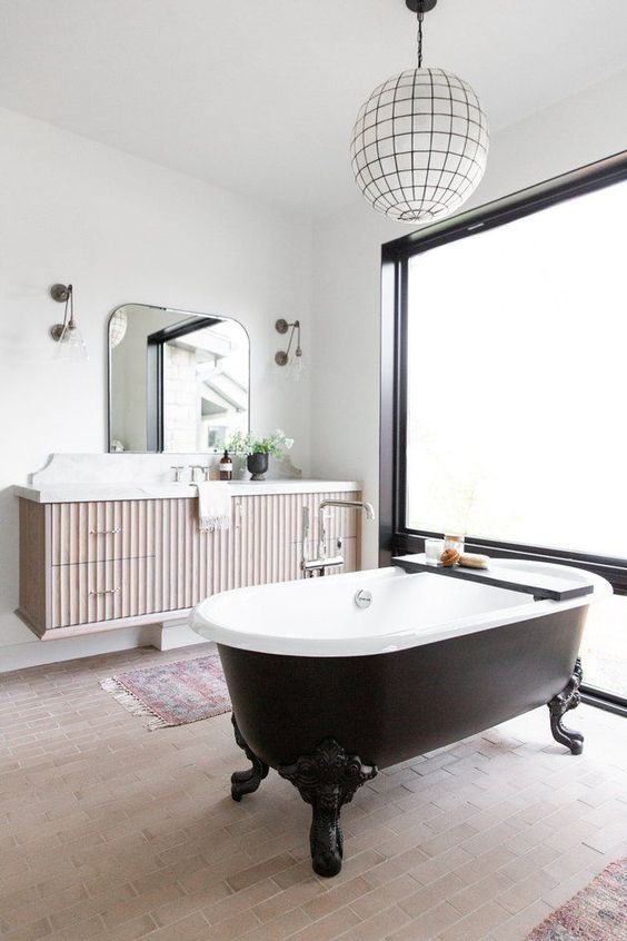 a fancy bathroom with white walls and a neutral tiled floor, a black vintage bathtub, a floating vanity and a pendant lamp plus a frosted glass wall