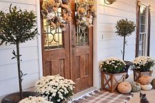a farmhouse fall porch with plaid rugs, potted blooms, heriloom pumpkins, potted trees and feather wreaths plus wall lanterns