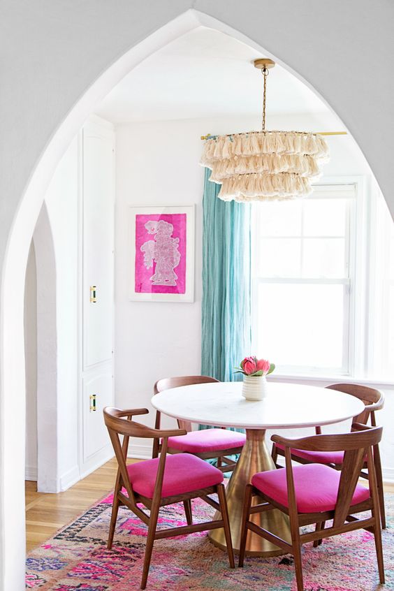 a lovely and airy dining space with a round table, hot pink chairs, a pink printed rug, a tassel chandelier and a hot pink artwork