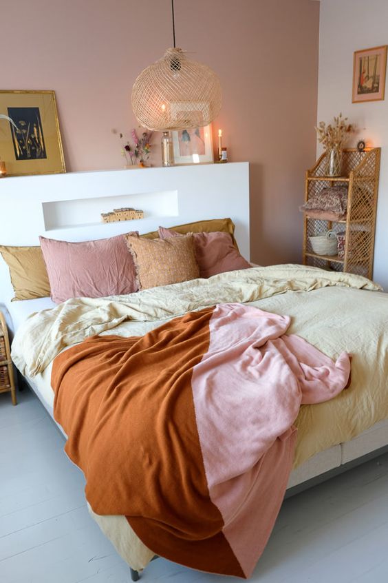 a lovely bedroom with a pink accent wall, a bed with an accent headboard, a pendant lamp and pretty bedding, a woven shelf and some artworks