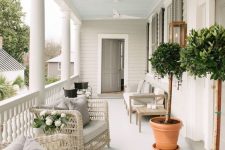 a lovely modern farmhouse porch with neutral wicker furniture, an upholstered bench and chairs, potted trees and lanterns