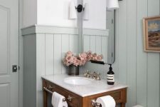 a modern country bathroom with pale aqua paneling, a stained vanity, white appliances, vintage lamps and some blooms