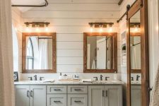 a modern country bathroom with white planked walls and a grey vanity, white subway tiles, mirrors in wooden frames with lights