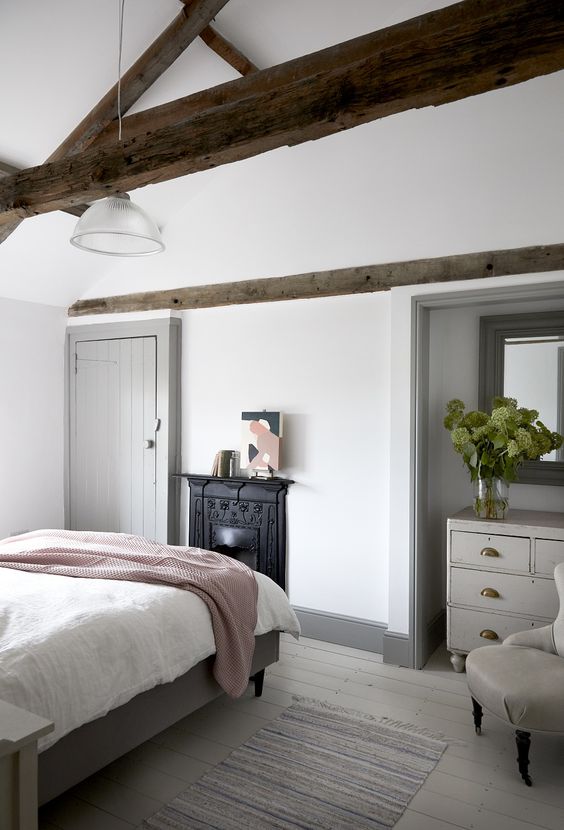 A modern country bedroom with dark stained wodoen beams, a non workign fireplace, neutral furniture and neutral textiles