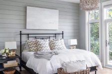 a modern country bedroom with grey planked walls, a black forged bed, neutral bedding, a wooden bead chandelier, a basket for storage