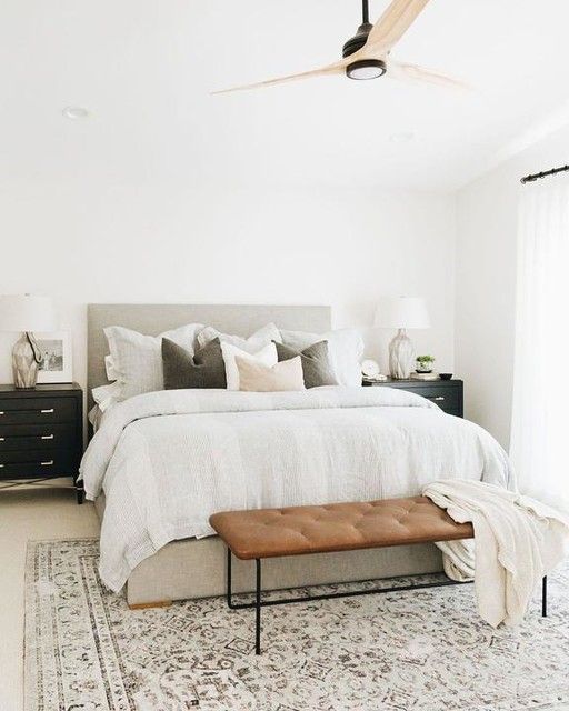 a modern country bedroom with neutral walls, a grey upholstered bed, a leather bench, black dresser nightstands and neutral textiles