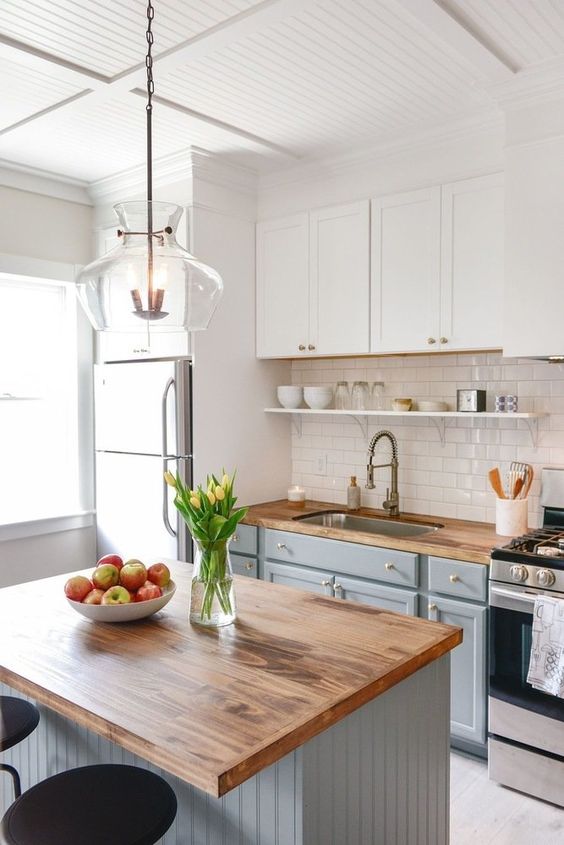 a modern country kitchen with white and light grey cabinets, butcherblock countertops, a shiny subway tile backsplash and a pendant lamp