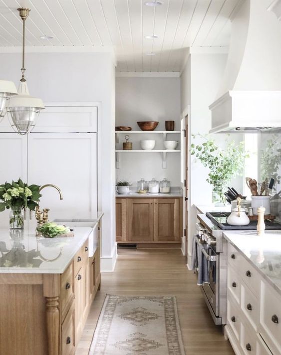 a modern country kitchen with white shaker style cabinets, a light stained kitchen island, white countertops and a large hood plus pendant lamps