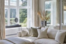 a modern country living room in neutrals, with mirrors, a creamy sectional, a bay window, a wooden table and cool textiles