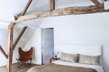 a modern farmhouse bedroom with stained wooden beams that highlight the attic ceiling, a neutral bed with a bench and a leather chair