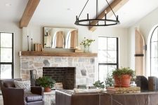 a modern farmhouse living room with a stone clad fireplace, wooden beams, leather furniture, a metal chandelier and potted plants